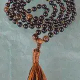 A Black Tourmaline and Tiger Eye Mala Beads Necklace with 108 beads plus 1 guru bead, handcrafted in the USA. Balances the Root and Solar Plexus Chakras, offering protection and courage. Suitable for meditation, spiritual practice, or as a bohemian accessory.