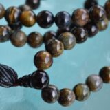 A handcrafted Black Tiger Eye Mala Necklace with 108 beads, designed for strength and protection, resonating with the Root and Solar Plexus Chakras and associated with Capricorn.